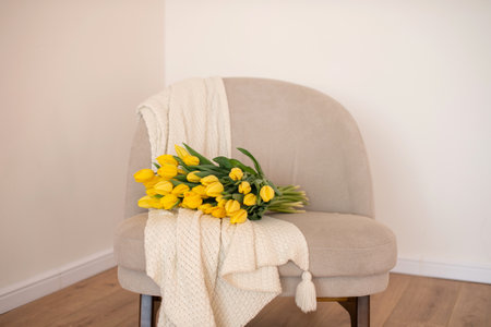 A bundle of bright yellow tulips rests on a cozy beige chair, draped with a knitted throw, inviting relaxation and reflectionの写真素材
