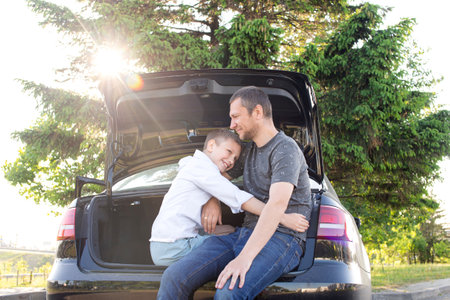 Father and son are sitting on the trunk of a car after a long road and laughing. Boy hugs dad, contour light, travelの写真素材
