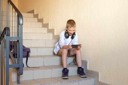 An angry boy spends time in the entrance on the stairs to play on the phone. Sit on the steps. Listen to music with headphones, look at the phone.の写真素材