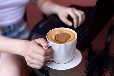 Close-up of a person's hand holding a coffee cup with heart-shaped foam, laptop in the backgroundの写真素材
