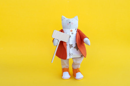 A soft toy cat in an orange jacket stands with a wooden sign with a place for text on a yellow background. photo of a toy standing on its feetの写真素材
