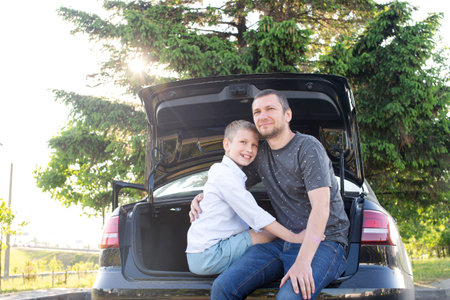 Dad and son are sitting in the trunk of a car at sunset. Hugging Dad and laughing. Relax after a long road, campingの写真素材