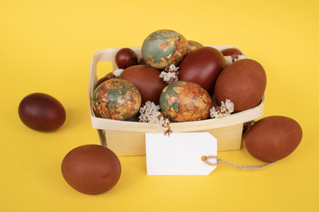 A vibrant display of Easter eggs naturally dyed in various hues, presented in a basket with a blank tag against a cheerful yellow backdropの写真素材