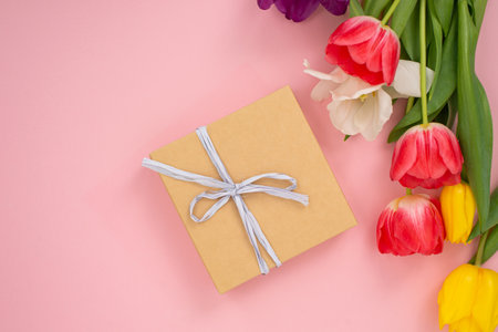 Kraft paper gift box with white ribbon next to vibrant tulips on a pink background. Flat lay arrangement for Mother's Day, spring holidays, and gift-giving concept for card, invitation designの写真素材