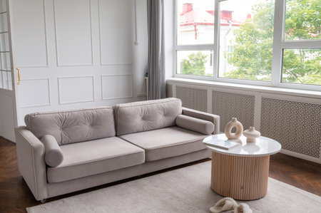 A chic living room boasts a comfortable beige sofa and a stylish round coffee table, with a view of greenery through a large windowの写真素材