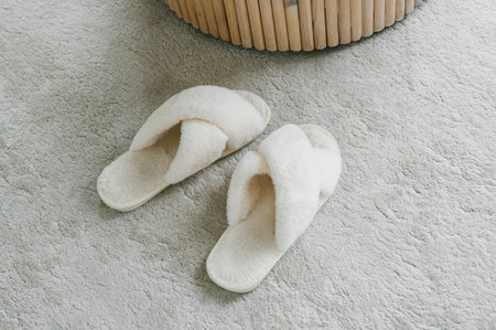 A pair of soft white slippers rests beside a stylish wicker basket, placed neatly on a textured plush carpet, evoking a sense of home comfortの写真素材
