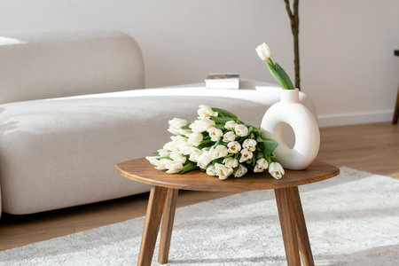 White tulips in modern vase on wooden table with sofa in the background. Interior design with spring flowers concept for poster, greeting card, home decorの写真素材