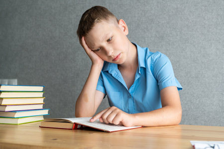 A young student focuses intently on reading a textbook among a stack of books, portraying dedication and education at homeの写真素材