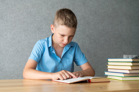 A focused young boy engrossed in reading a book at a wooden desk, with a colorful stack of books beside him, symbolizing education and learningの写真素材