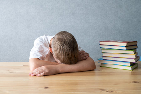 A tired young student rests his head on arms beside a stack of colorful books, symbolizing academic fatigueの写真素材