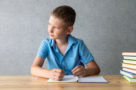 A preteen boy wearing a blue shirt is deeply focused on reading from a book with a stack of colorful books beside himの写真素材