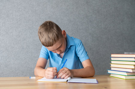 A focused young boy in a blue shirt diligently writes his homework in a notebook, sitting at a wooden desk besides colorful textbooksの写真素材