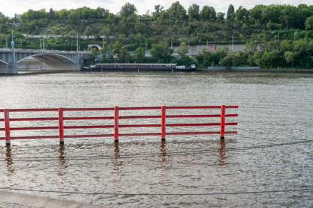 Red railing partially submerged in water by a riverside with a bridge and greenery in the background. Outdoor sceneの写真素材