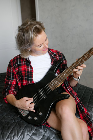 A middle-aged woman in a plaid shirt comfortably plays a black electric guitar while sitting on a bed, showcasing musical leisure activity at homeの写真素材