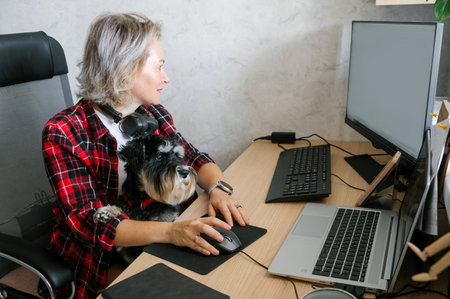 A smiling woman in a checkered shirt works on a computer at home, enjoying the company of her adorable black dogの写真素材