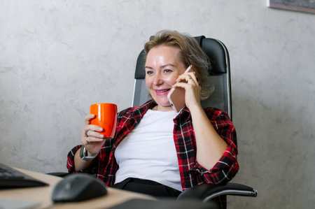 Capture a slice of office life with this image of a cheerful woman holding her vibrant orange mug while chatting on the phone at her deskの写真素材