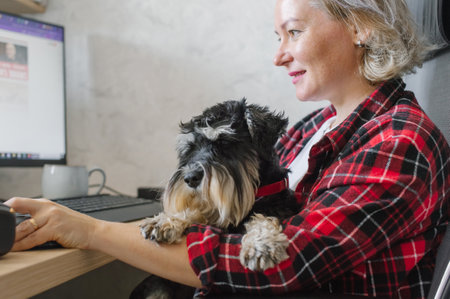An elderly woman in a plaid shirt enjoys the company of her small dog while focusing on her computer work in a cozy home office settingの写真素材