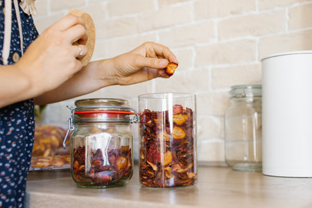 A close-up view of unidentifiable woman's hands arranging dried fruit in airtight glass jars, showing healthy snacking optionsの写真素材