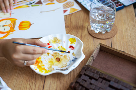 Close-up of an artist's hands as they intricately paint vibrant fall-themed illustrations,...