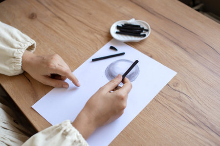 Close-up view of hands skillfully drawing with charcoal on a blank paper, next to art supplies on a wooden surfaceの写真素材