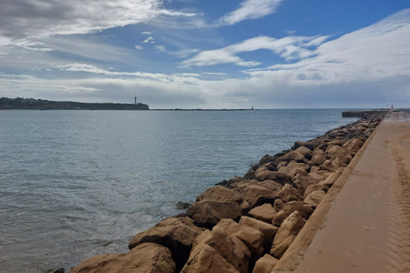 Capture the tranquil beauty of a coastal scene, featuring a lighthouse in the distance under a softly clouded skyの写真素材