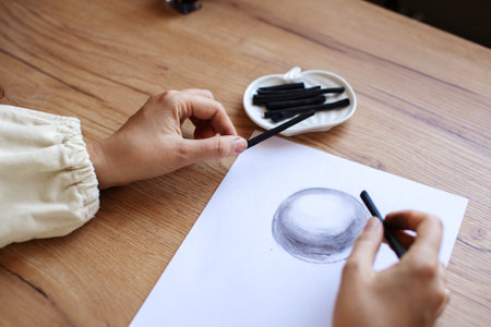 A close-up view of an artist's hands as they skilfully draw a shaded sphere on a white paper using charcoalの写真素材
