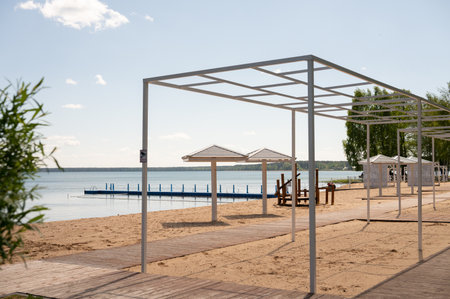 Empty beach with white pergolas and wooden path by tranquil lake under clear blue sky.の写真素材