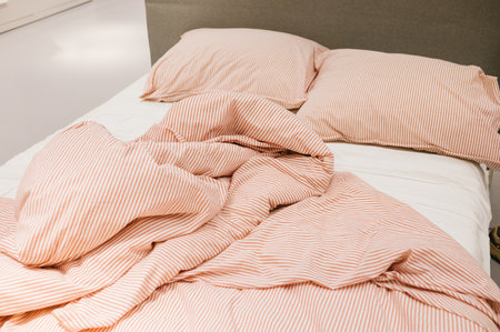 Cozy unmade bed with striped pink duvet and pillows in sunny bedroom scene.の写真素材