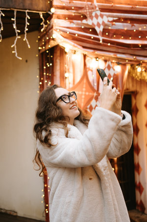 Young caucasian female capturing festive moments with smartphone under twinkling lights.の写真素材