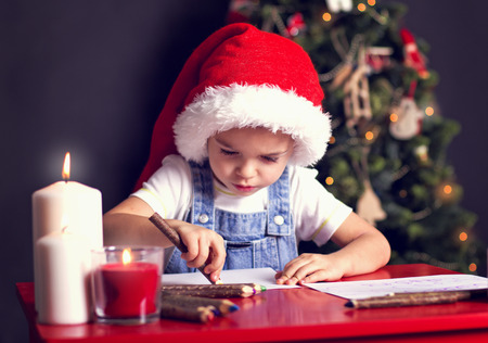 Christmas boy in the red hat writing letter to Santa on red table with candles and christmas theeの写真素材