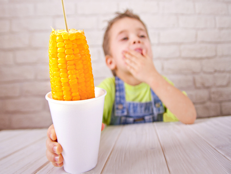 Cute boy holdind boiled sweet corn in cup on white bricks wall background.