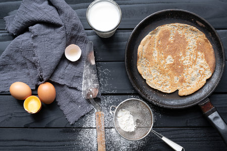 Hot pancake in black pan on black table with flour, milk and eggs.の写真素材