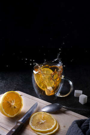 falling slices of lemons into a mug with hot tea on black background, tea splashes in different...
