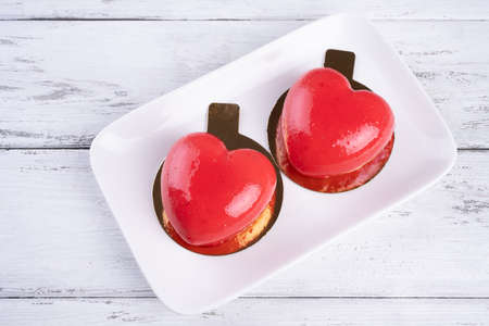 Two mousse cakes in the form of a red heart on a white plate on white wooden background
