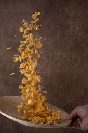 dry cornflakes falling into wooden bowl on brown background. Levitating food