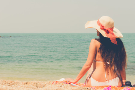 Beautiful asian bikini woman sitting on beach  looking at  the sea with white large hat vintage retro filter effect for exotic holiday romance relaxing vacation conceptの写真素材