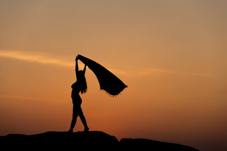 Silhouette of half naked confident woman man doing a freedom pose against the sun on rocky ground on a sun set sky near beach for wining achievement, freedom, overcome, result performance conceptの写真素材