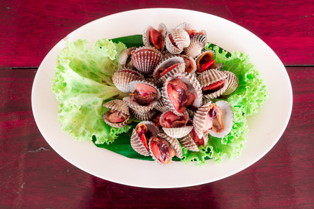 Authentic half boil cockle salad cooked thai style on white plate and red wooden table. Exotic food concepts.の写真素材