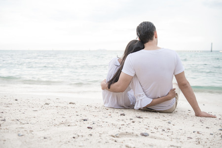 Couple holding each other on beach. Young happy interracial couple on beach holding each other sitting down. Asian woman, Caucasian man. Young mixed race romance concept.の写真素材
