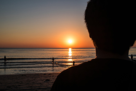 Silhouette, blurred close-up rear view of a man standing viewing sunset at the beach, selective focus on horizon and the sunの写真素材