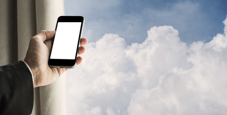 Businessman using mobile phone with curtain opened, and blue sky with fluffy clouds backgroundの写真素材