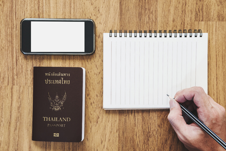 Hand holding pencil writing notebook and mobile phone with Thai passport on wooden desk, vintage toneの写真素材