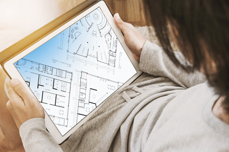 Asian guy using digital tablet in living room, with architectural layout plan on screen