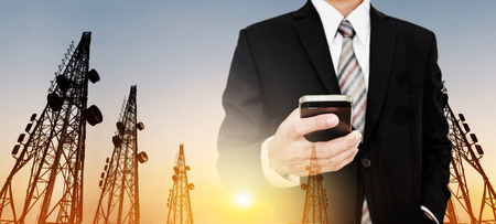 Panoramic, Businessman using mobile phone with Telecommunication towers with TV antennas and satellite dish in sunsetの写真素材