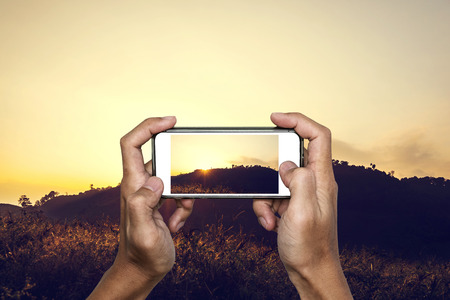 Hand using smartphone taking photo of tropical mountain landscape in sunsetの写真素材
