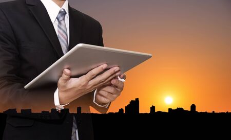 Businessman working on digital tablet, with silhouette buildings in the city in sunset backgroundの写真素材