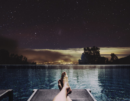 Relaxing in holidays, a man feet on bed at swimming pool at night with city lights and stars on sky backgroundの写真素材