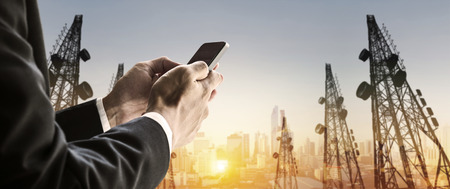 Businessman using smart phone with blurred defocus double exposure panoramic cityscape and telecommunication towers in sunriseの写真素材