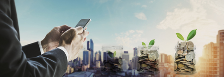 Businessman using smartphone with coins in bottles, and sunrise in the city backgroundの写真素材