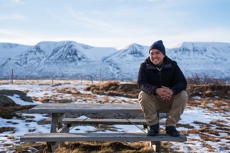 Asian guy sitting on wooden bench with smiling face, traveling in winterの写真素材